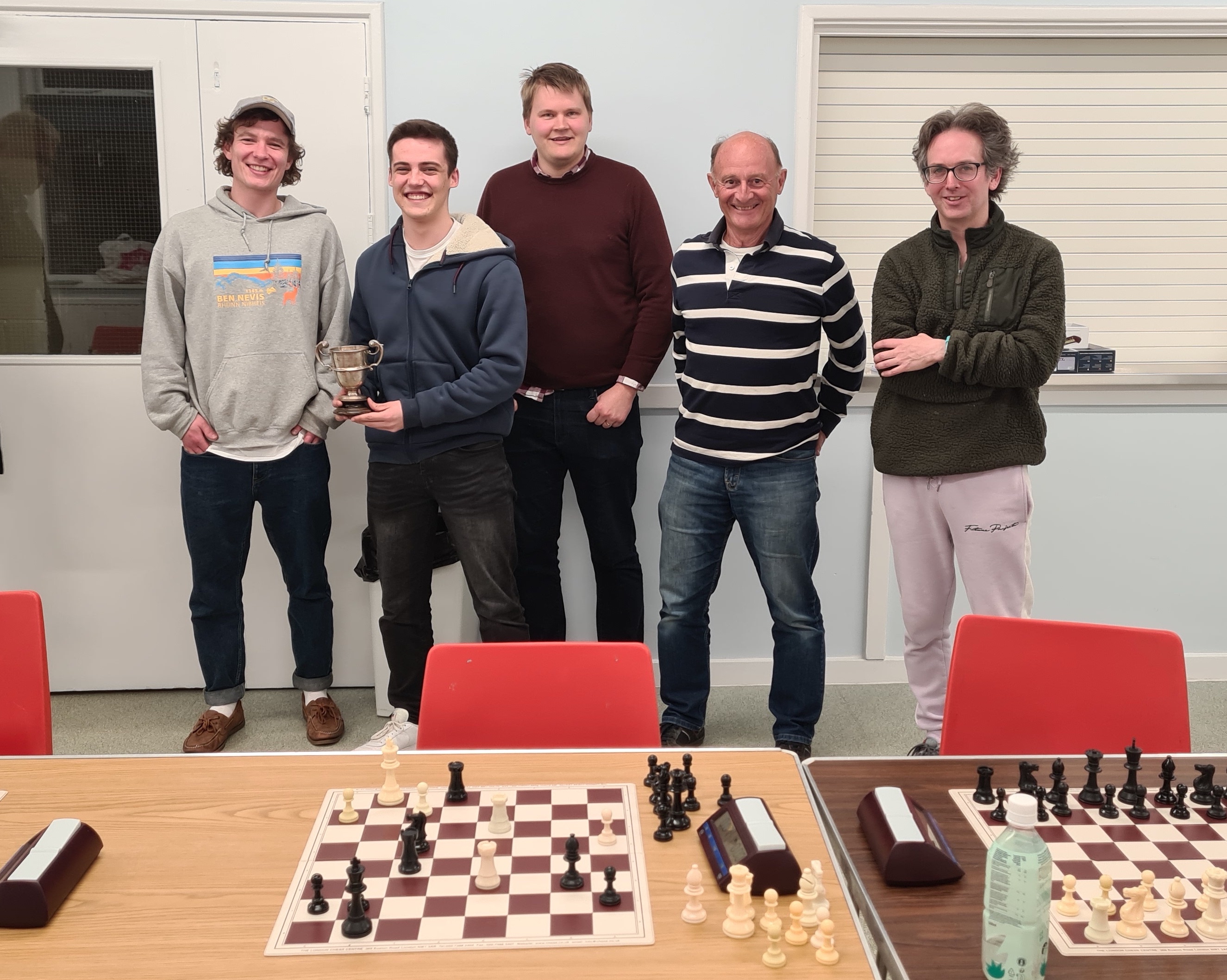 5 chess players posing for a photo behind the boards, the second player holds a trophy